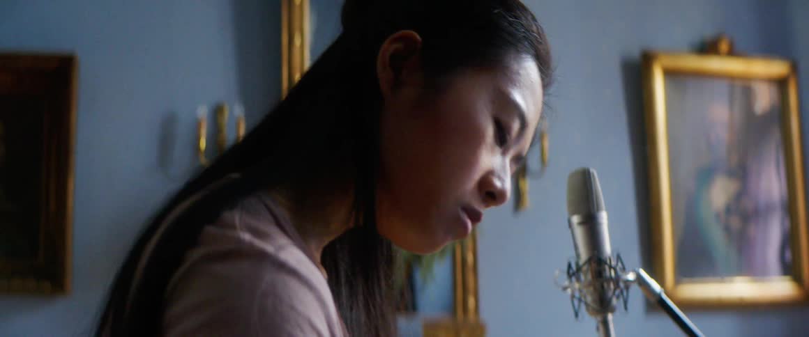 A woman sitting at a desk writing on a paper next to a laptop with a microphone a wall and picture frames in the background