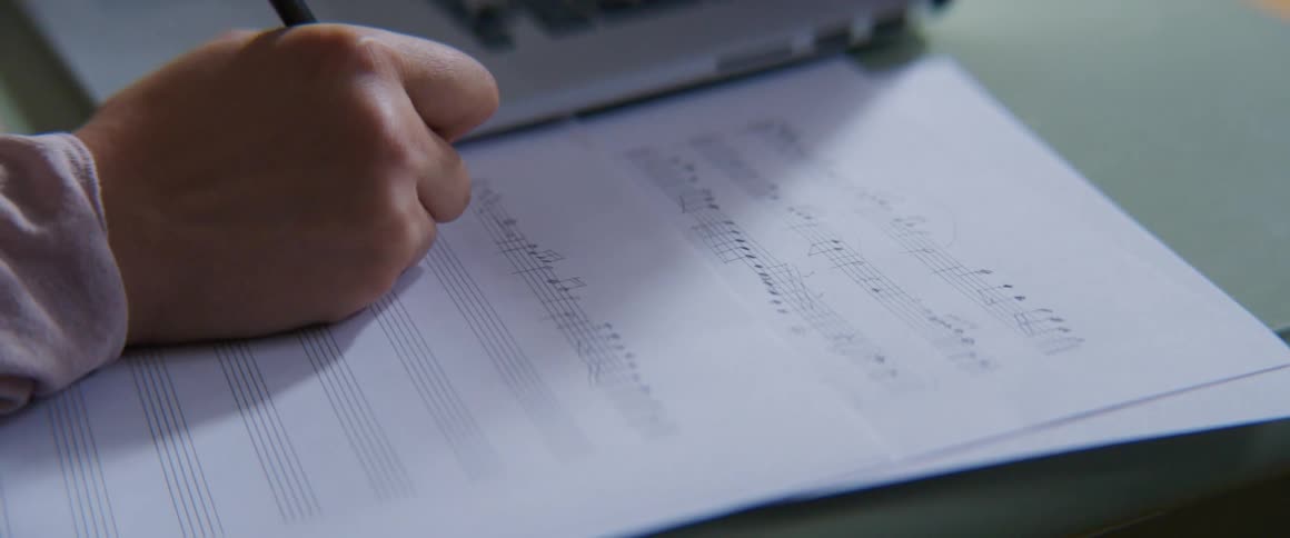 A musician writing music on a music sheet next to a laptop