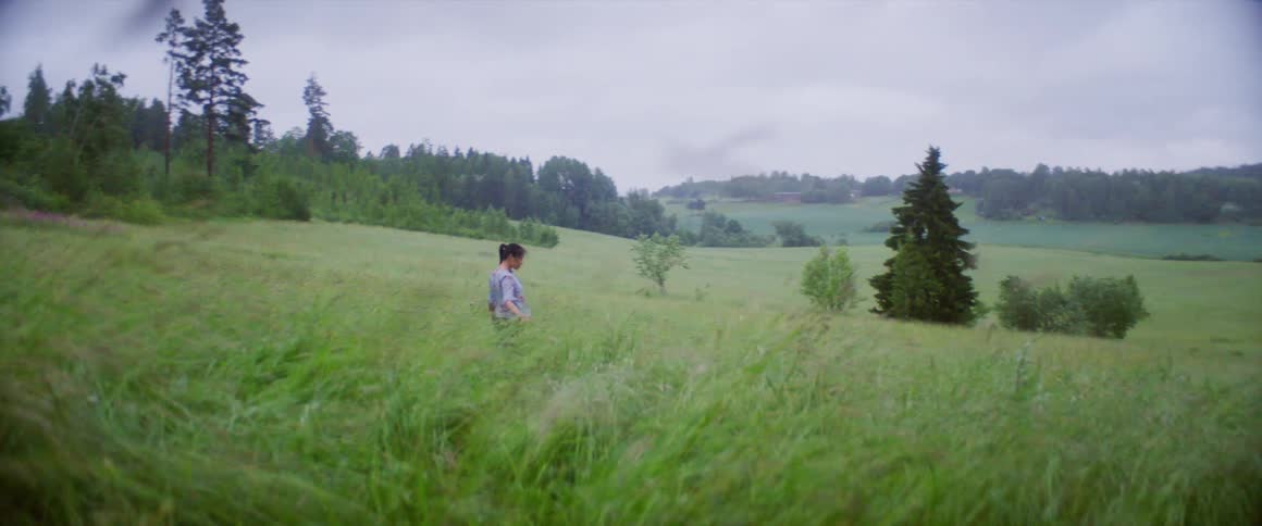 Woman walks through grass bordered by hills and forest