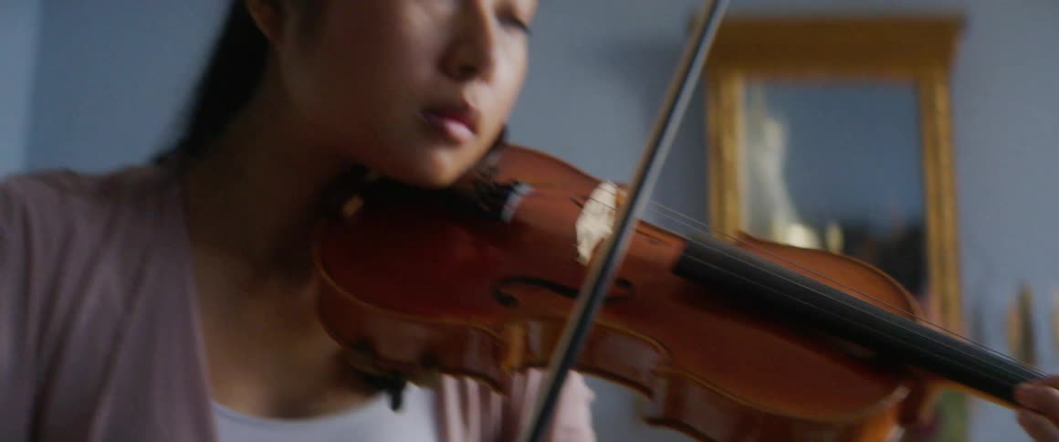 Asian woman holding a violin against her neck and moving a bow plays a violin in a room with paintings