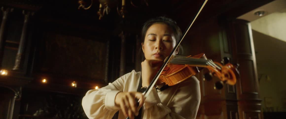 Asian woman holding a violin against her neck and moving a bow plays a violin under a chandelier in a large room