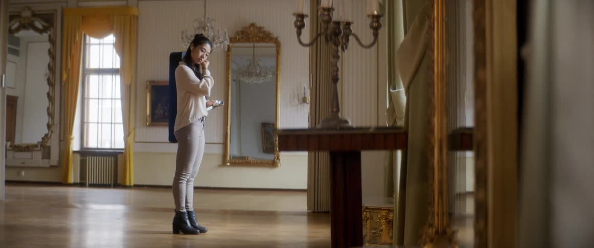 A young asian woman carrying an instrument case on her back places ear buds into her ear while holding a smartphone in a large room in a house during the day