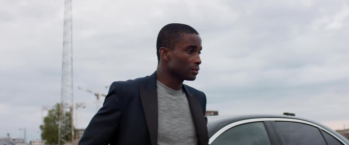 Man wearing suit jacket stands near car under cloudy sky