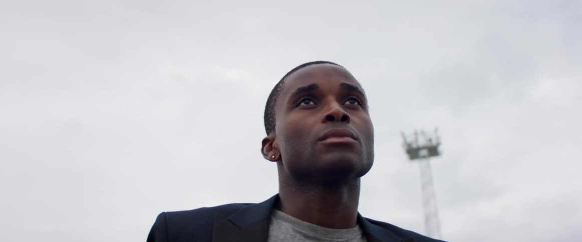 A man with a blazer standing outside with the sky and clouds in the background
