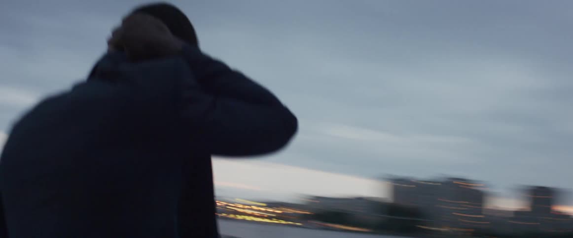 Man rubs his head looking around as city lights shine form skyline across water