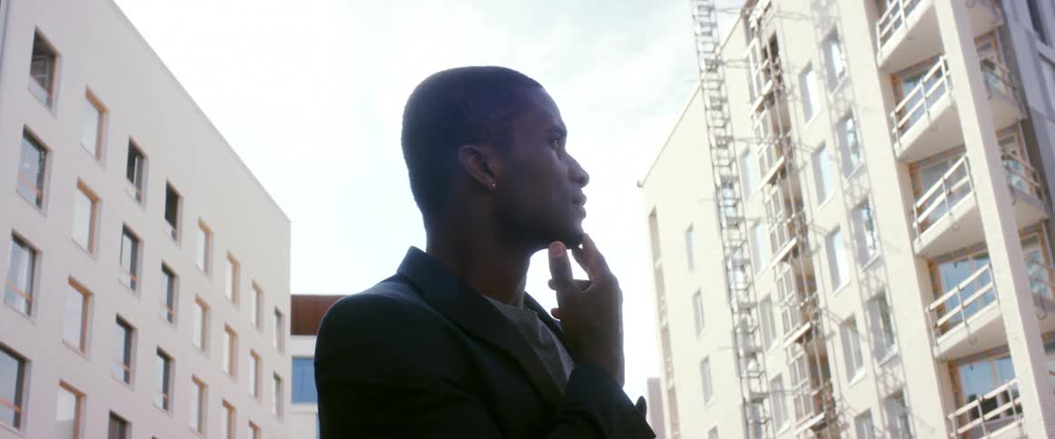 A man stands outside of large city buildings tapping his fingers on his chin in thought