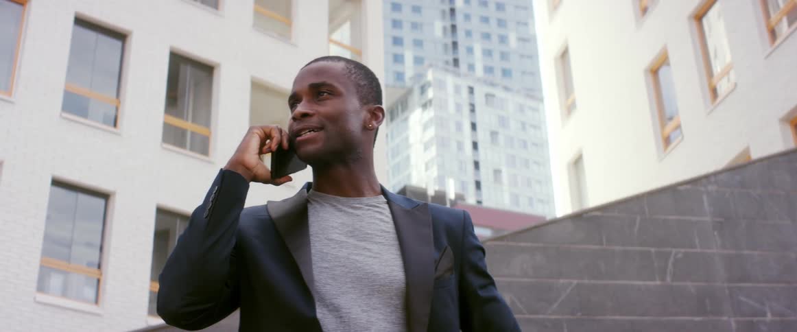 A man standing outdoors in a metropolis during the day gestures with his hands while talking on a smartphone
