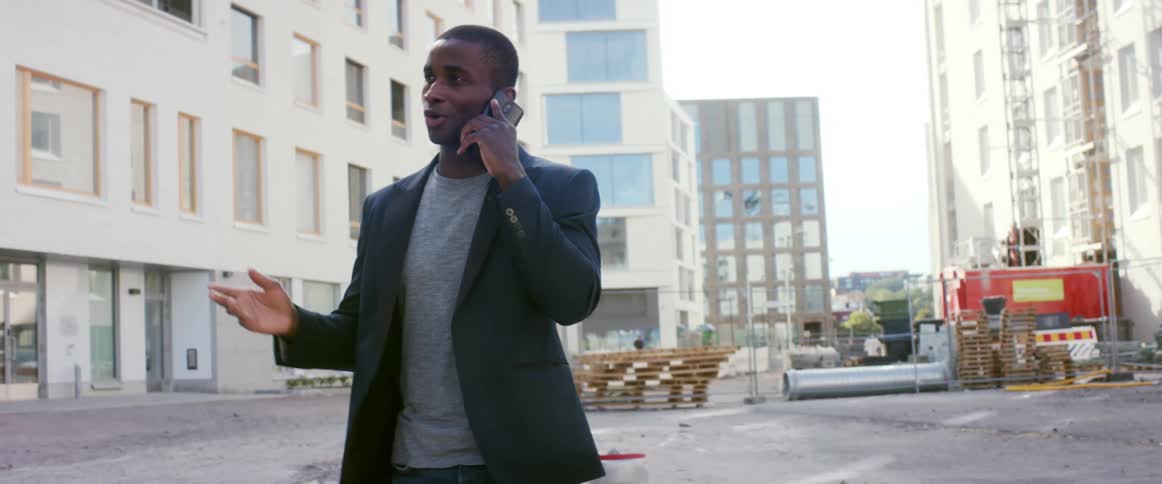 A man with a blazer and a smart phone standing in a city with buildings the sky and clouds in the background
