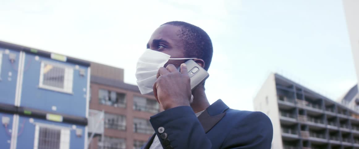 A man wearing a medical mask talks on his smartphone while gazing around at the buildings surrounding him