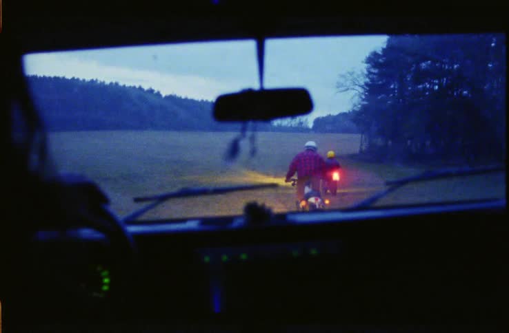 A person driving a vehicle on a field and two people on motorbikes come out of the forest and cut in front and drive off