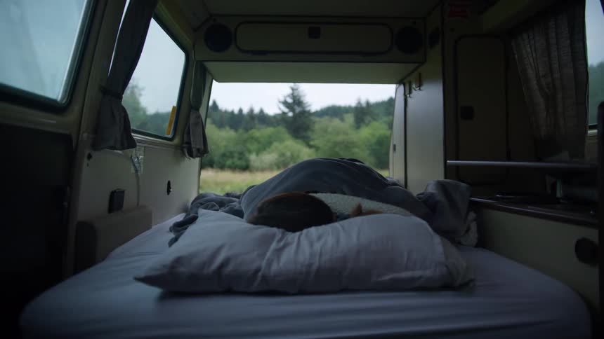 Woman sleeps on bed in a vehicle with doors open and nature outside