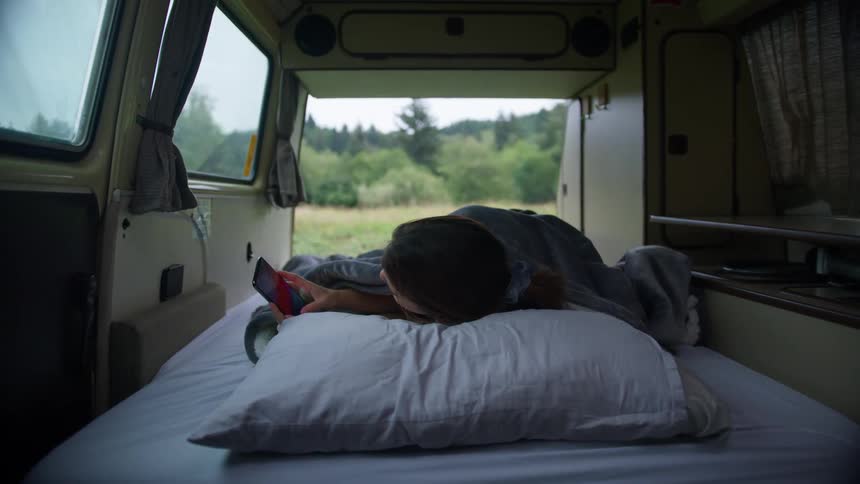 Woman wakes up to check mobile phone then leans while looking out open caravan door toward hill