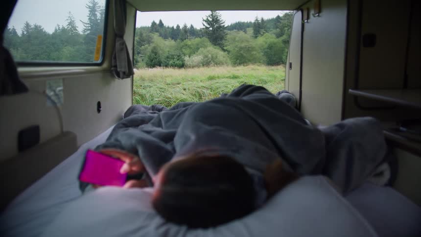 Person lays on bed inside covered in a blanket van playing on phone there is a wooded area outside of the windows and door that you can see in dim sunlight past a clearing with tall grass
