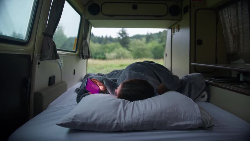 A female lays on a makeshift bed inside a van looking at her cell phone