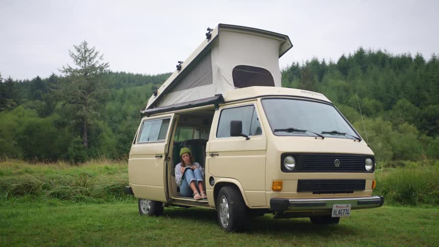 A woman sits in an old camper van outside in a rural setting