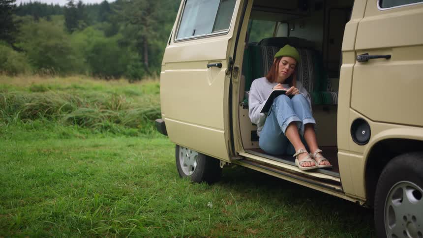 A woman sits in an open van in a rural setting writing in a notebook