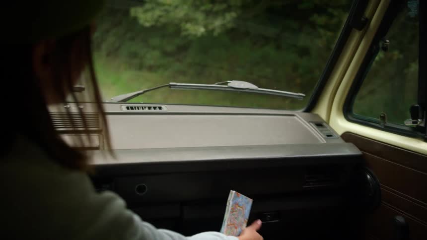 Woman sitting in parked vehicle on side of forest road consults map from glove compartment
