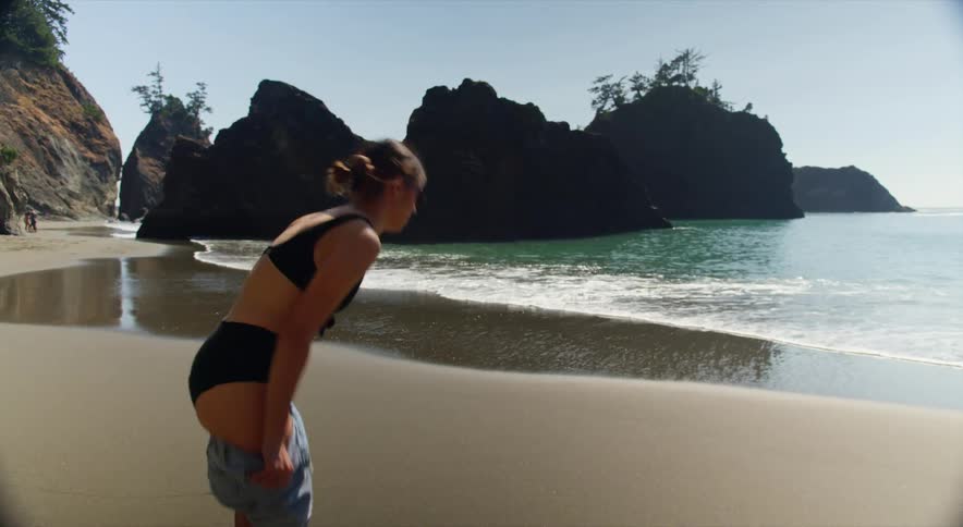 A woman on the beach walks closer to the ocean and takes off her denim shorts