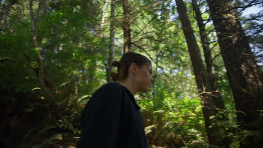 Woman in shirt pants and a sweater walks down a forest trail while strands of hair that fell out of her pony tail swing in her face in the shade of big trees