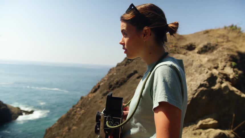 A woman with a camera and sunglasses standing near the coast with the ocean the sky and clouds in the background