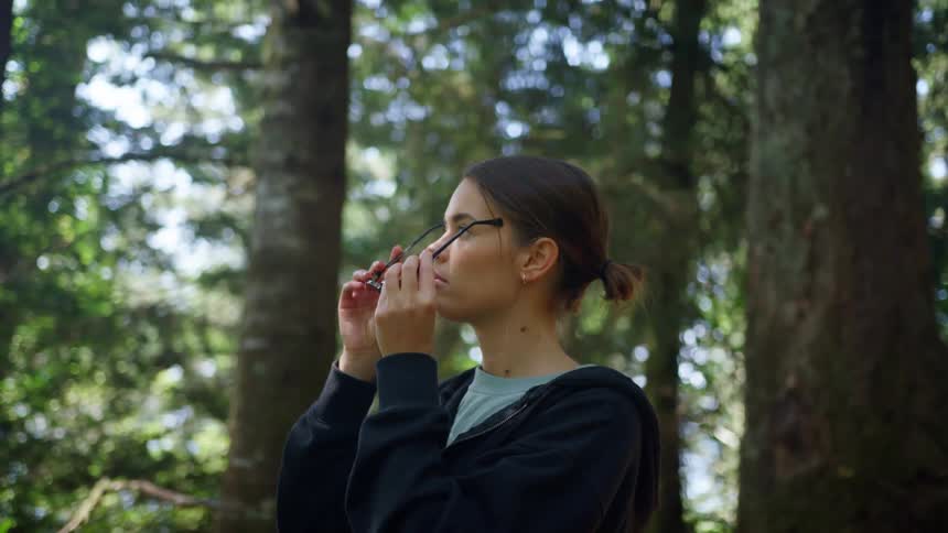 Woman with a black zip hoodie puts on sunglasses in the woods