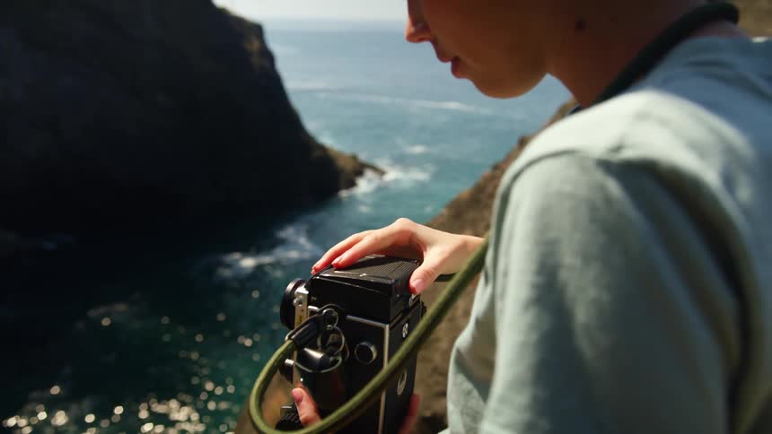Woman stands on ocean side cliff getting the camera she has on a strap around her neck ready to take a picture of tress on another cliff