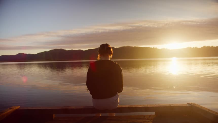 Woman sits on dock looking toward forest on horizon as rising sun reflects off rippling water