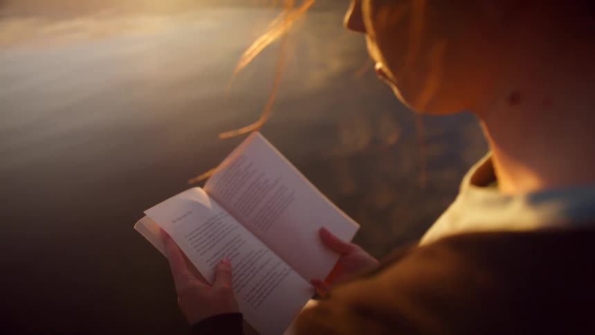 A young woman standing on the shore reading a book