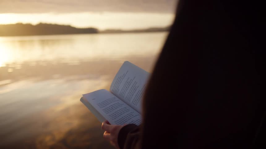 Woman wearing a dark sweater and light pants sits by a lake reading a book at sunrise
