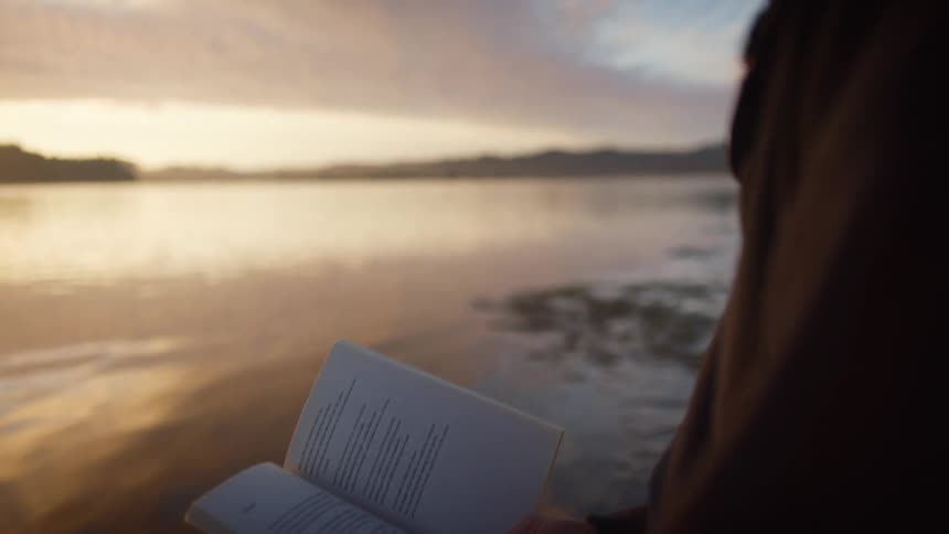 A woman is reading a book by the beach at dusk