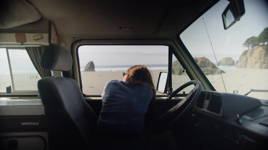 A woman rests her head on the open window of a van parked on a beach during the day