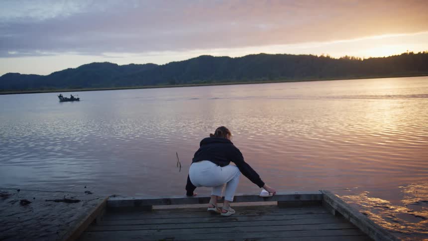 A woman approaches the shoreline holding a book and sits down on a dock with the sun low in the sky
