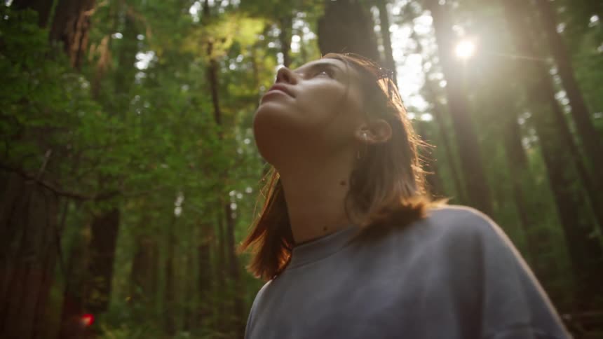 A woman with sunglasses standing in a forest with trees the sky and clouds in the background