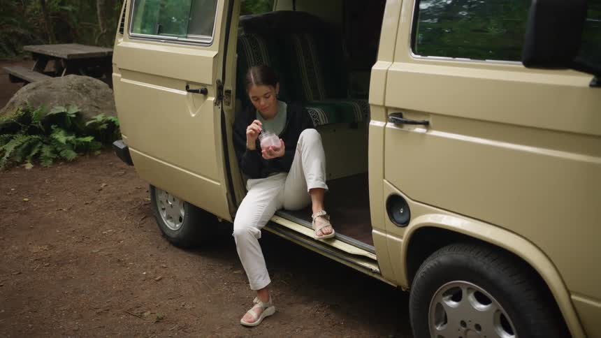 A woman sitting in a vehicle eating yogurt with a bench and trees in the background