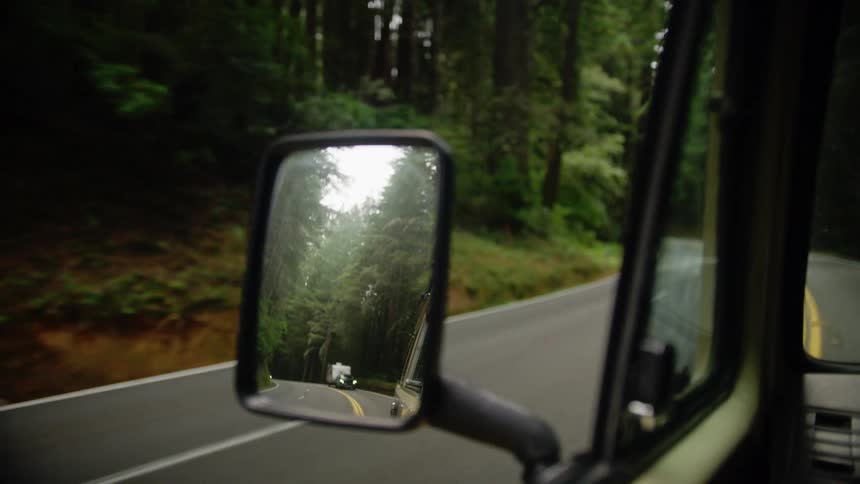A vehicle with a rearview mirror driving down a road surrounded by trees with the sky and clouds in the background