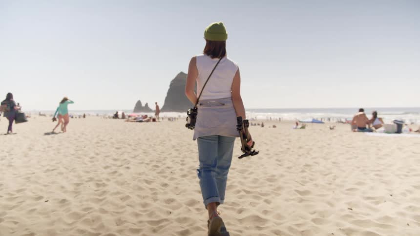 Photographer walks across beach sand carrying camera equipment toward ocean and other beach goers