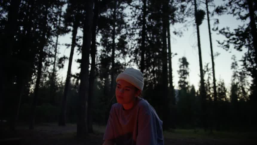 Woman with a white beanie and a sweatshirt sits by a campfire in the forest at dusk