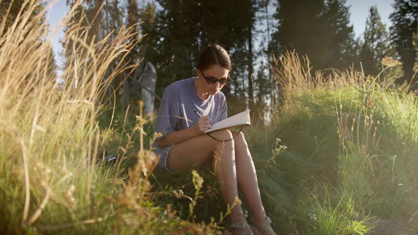 Woman wearing sunglasses sits on ground near conifer woods writing in journal