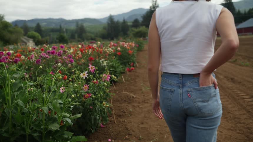Woman walks past flower garden on dirt road covered in tractor tracks with farm buildings and mountains in distance