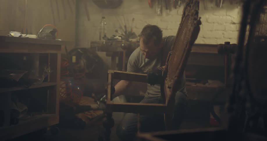 A man uses a tool on a broken chair inside a dim workshop