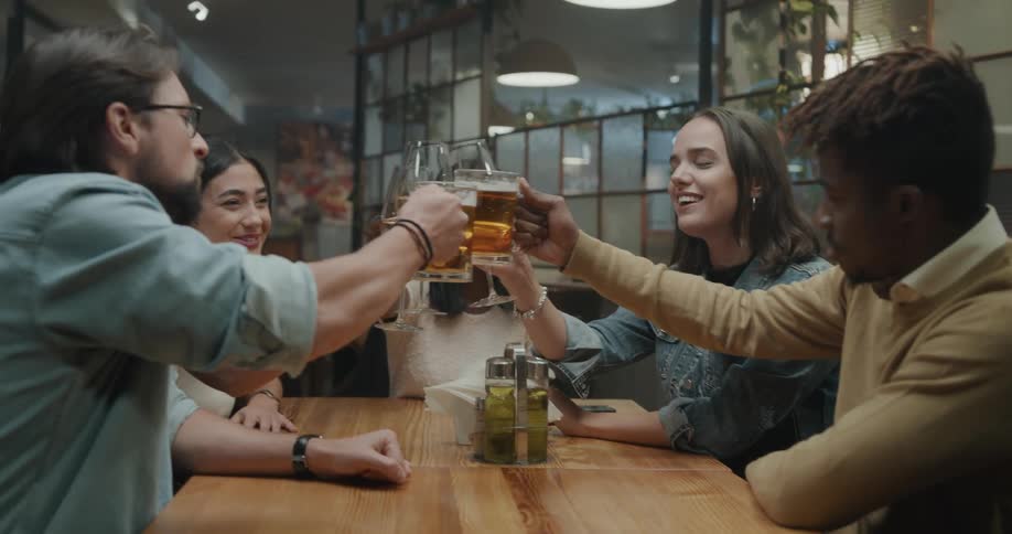 A group of young adults sit at a restaurant table and cheers their wine and beer glasses