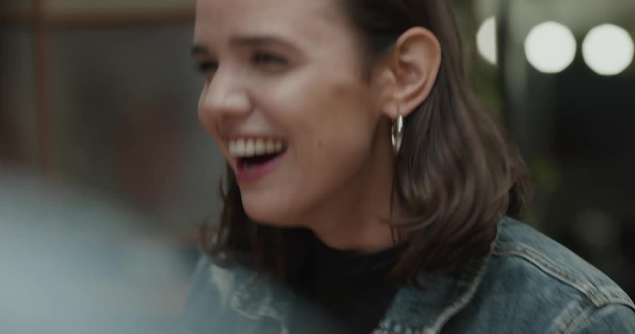 Woman with hoop earrings and a denim jacket sits at at table and laugh with her friends in a restaurant
