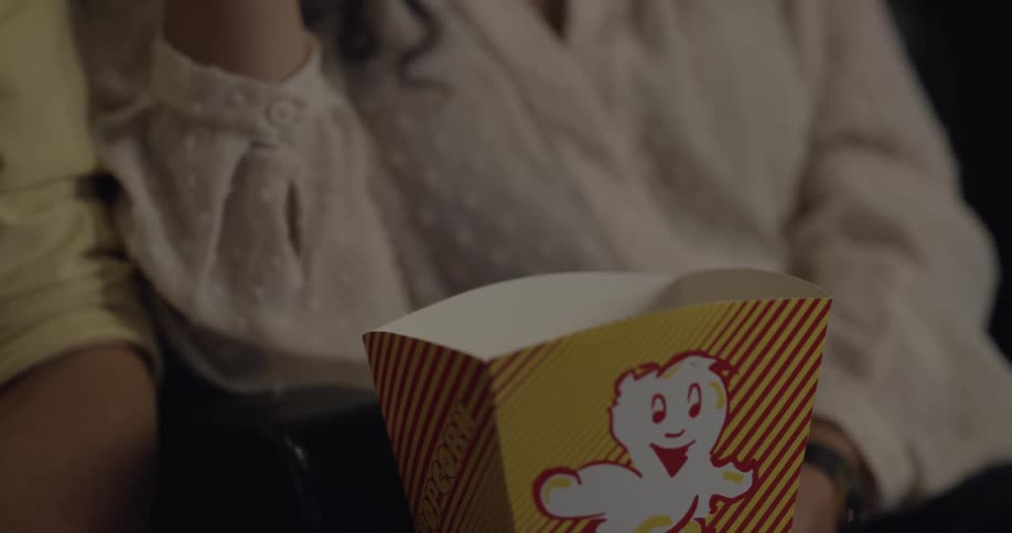 A man and woman eat popcorn from a cardboard container while sitting in next to each other in a theater