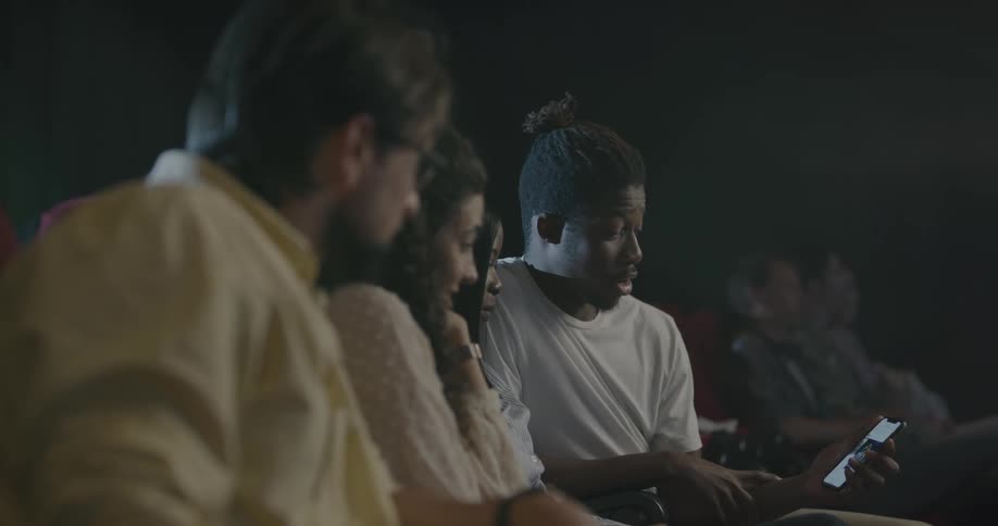 People sitting in a movie theater laugh as they look at a mans cell phone