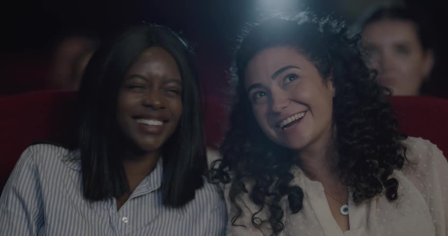 Two female friends talk to each other while watching a movie in the movie theater