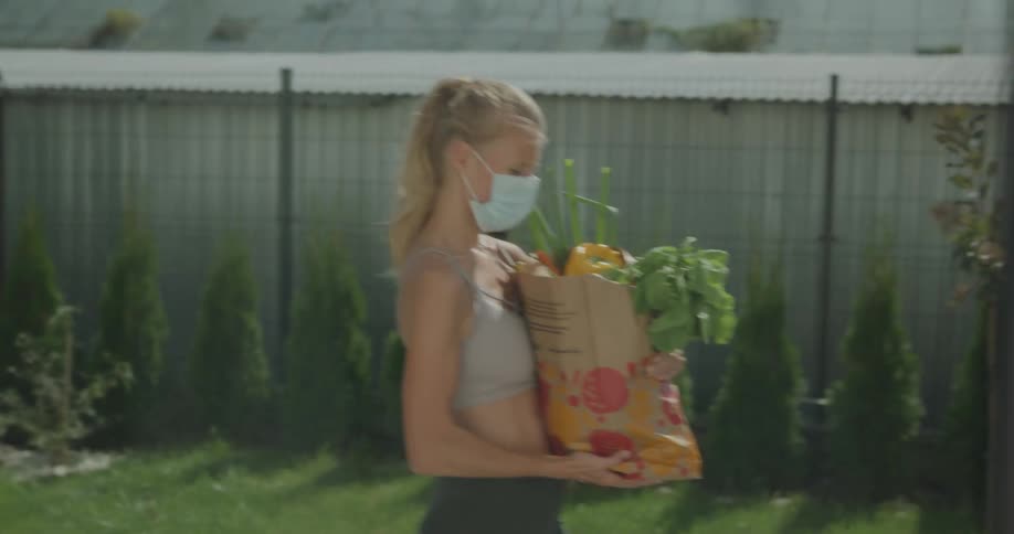 A blonde woman wearing a face mask carries a bag of groceries across a yard