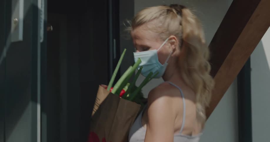 A woman wearing a mask carries a bag of groceries in to her home