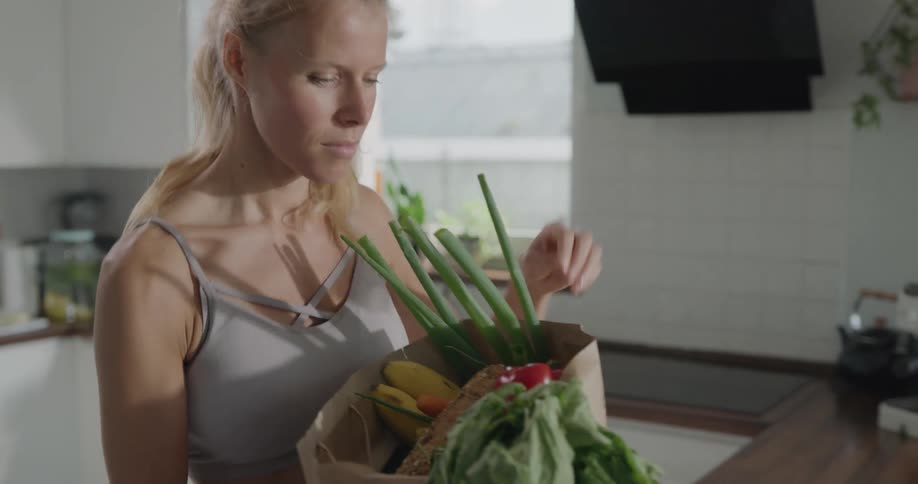 Blonde woman with a ponytail takes off mask and takes out the vegetables from the bag in the kitchen