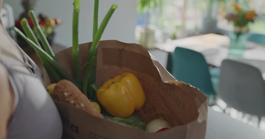 A woman pulls out vegetables from a paper shopping bag and places them on a kitchen counter