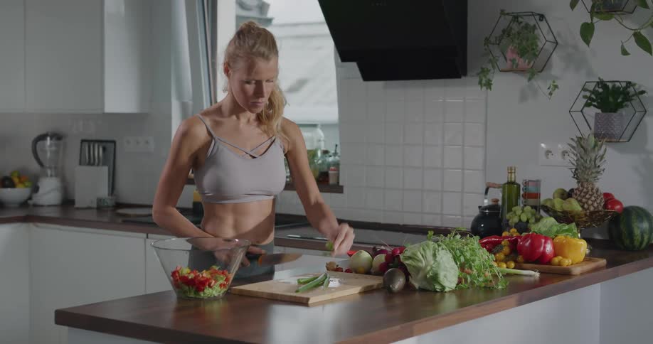 Blonde woman wearing exercise gear stands at kitchen counter slicing vegetables for a salad